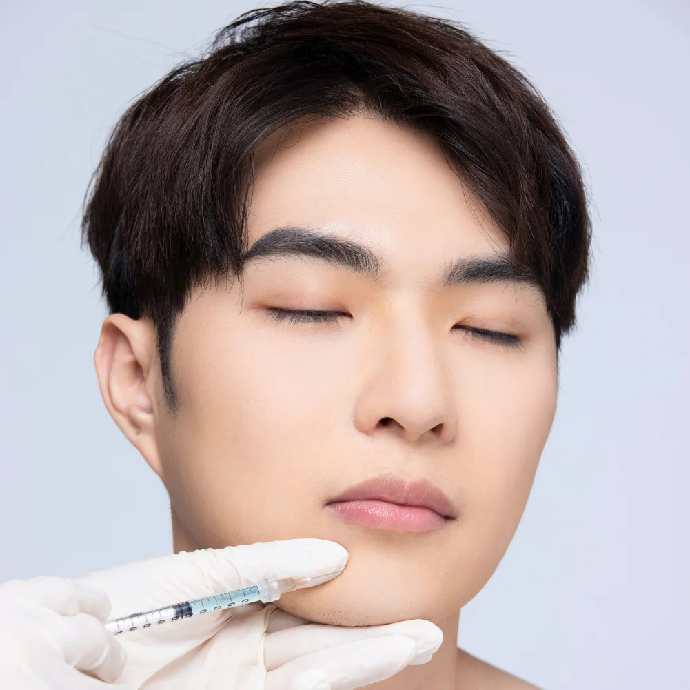 Young adult prepared for facial dermal filler injection, syringe held near lower cheek.