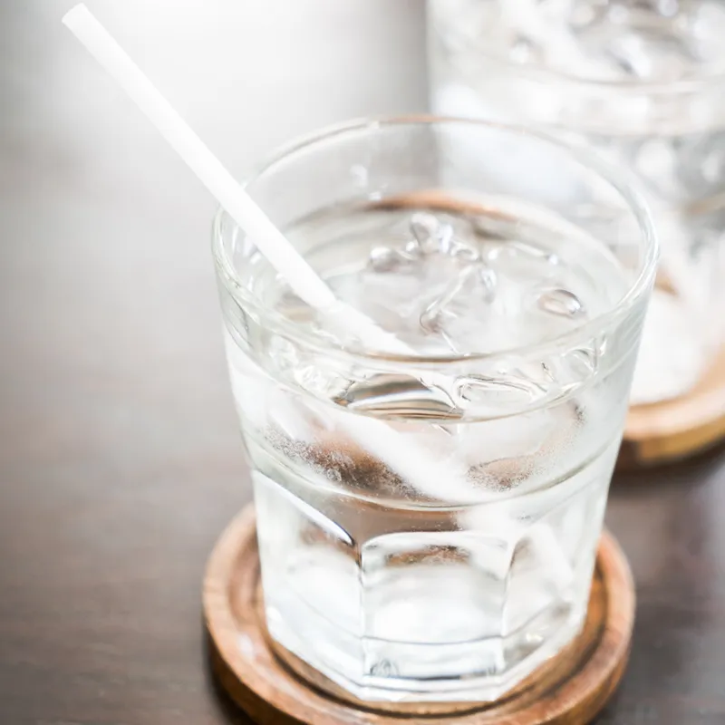 Close-up glass of ice water with straw on wooden coaster, condensation and melting ice