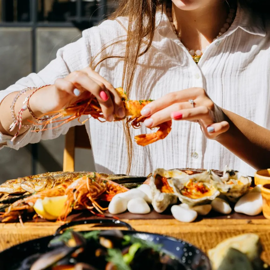 Sunlit hands peeling prawn over rustic seafood platter with oysters, mussels, and lemon wedges