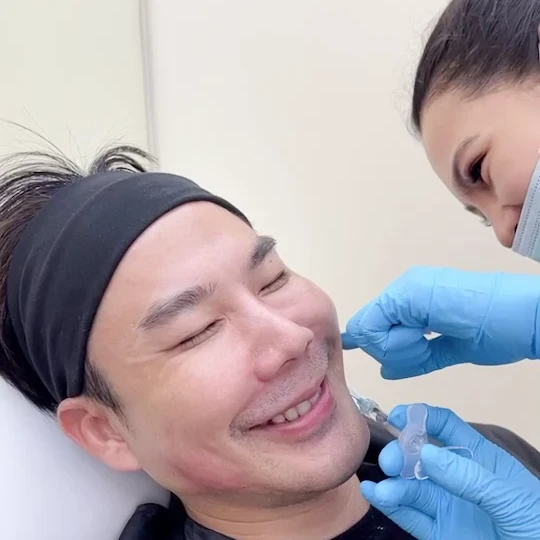 Smiling male patient receiving cheek filler treatment from masked, gloved practitioner in clinic