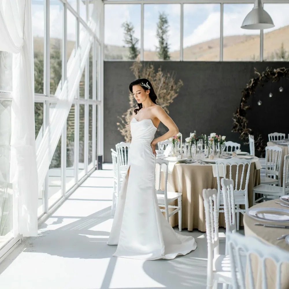 Sunlit bride in strapless white gown at wedding venue with reception tables and floral arch