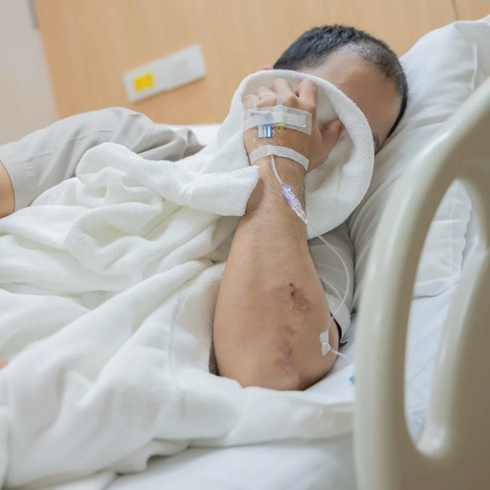 Patient in hospital bed with IV line taped to hand, resting under white blanket
