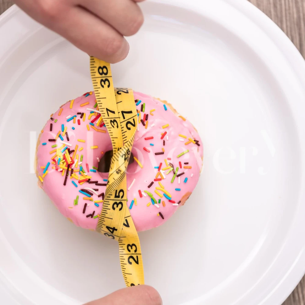 Pink frosted donut with sprinkles wrapped by measuring tape on white plate, portion control concept