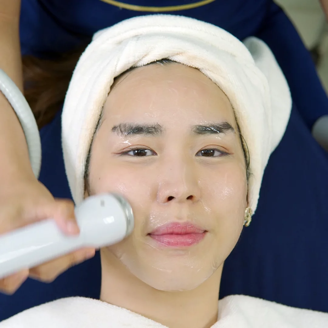 Towel-turbaned client receiving spa facial with serum, eye patches, and esthetician device
